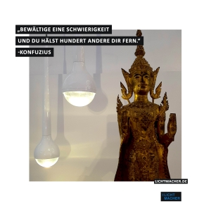 Zwei tropfenförmige Leuchten, neben einer goldenen Statue von einem indischen Budda. Dazu der Spruch "Bewältige eine Schwierigkeit und due hälst hunderte andere dir fern." von Konfuzius